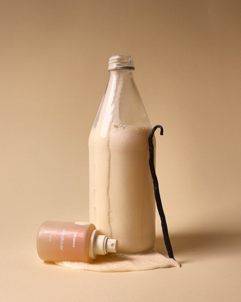 A perfume bottle resting on its side beside a milk bottle and a vanilla pod, in warm skin-toned light